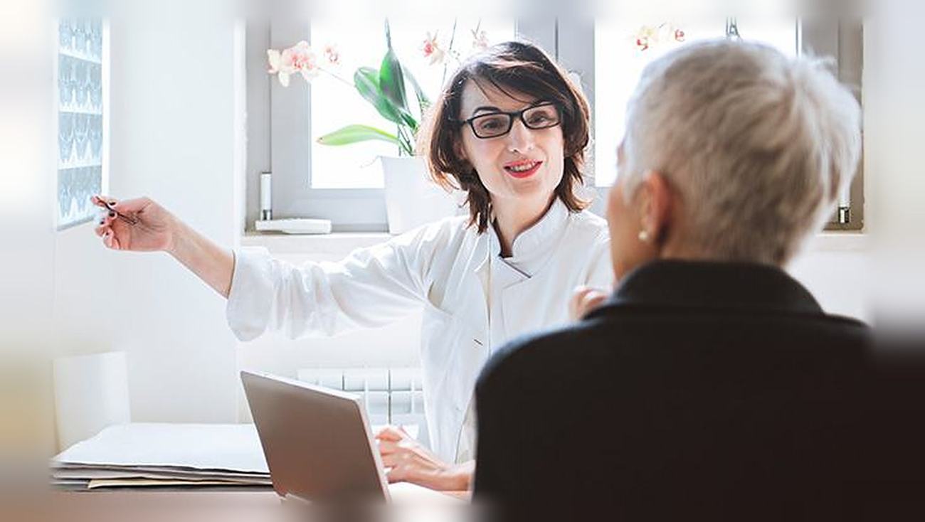 A smiling health professional in a white coat talks with an older person across a desk.