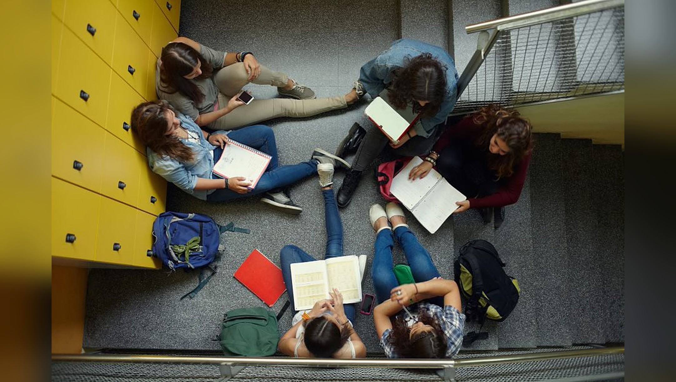 Six secondary students sit in a circle on a school stairwell landing beside yellow lockers, reading notebooks together.